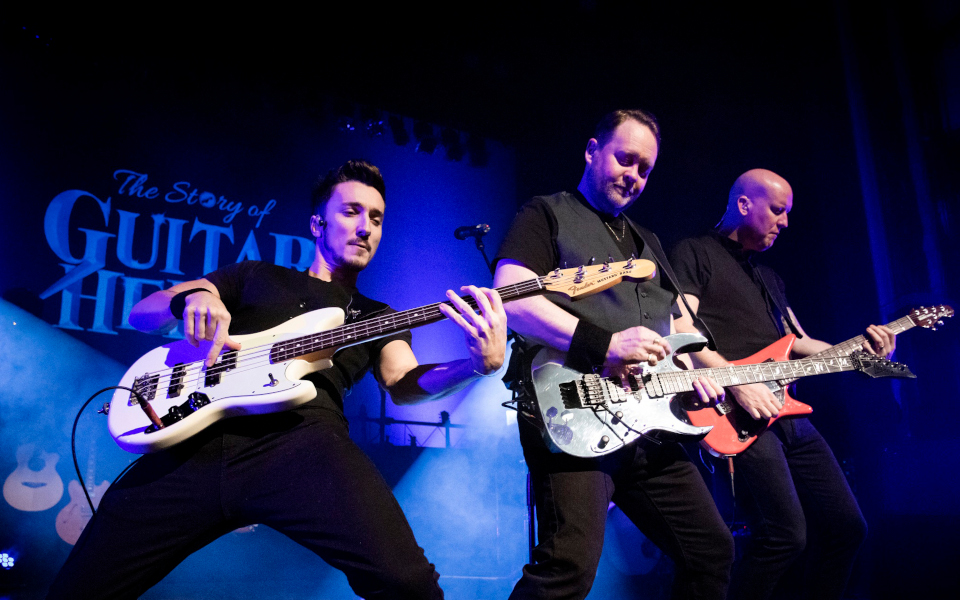 The Story of Guitar Heroes Three guitarists playing electric guitars on stage