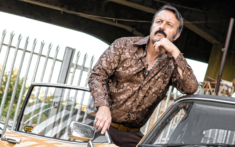 Mike Bubbins Comedian Mike Bubbins wearing a patterned shirt and leaning on a vintage car.