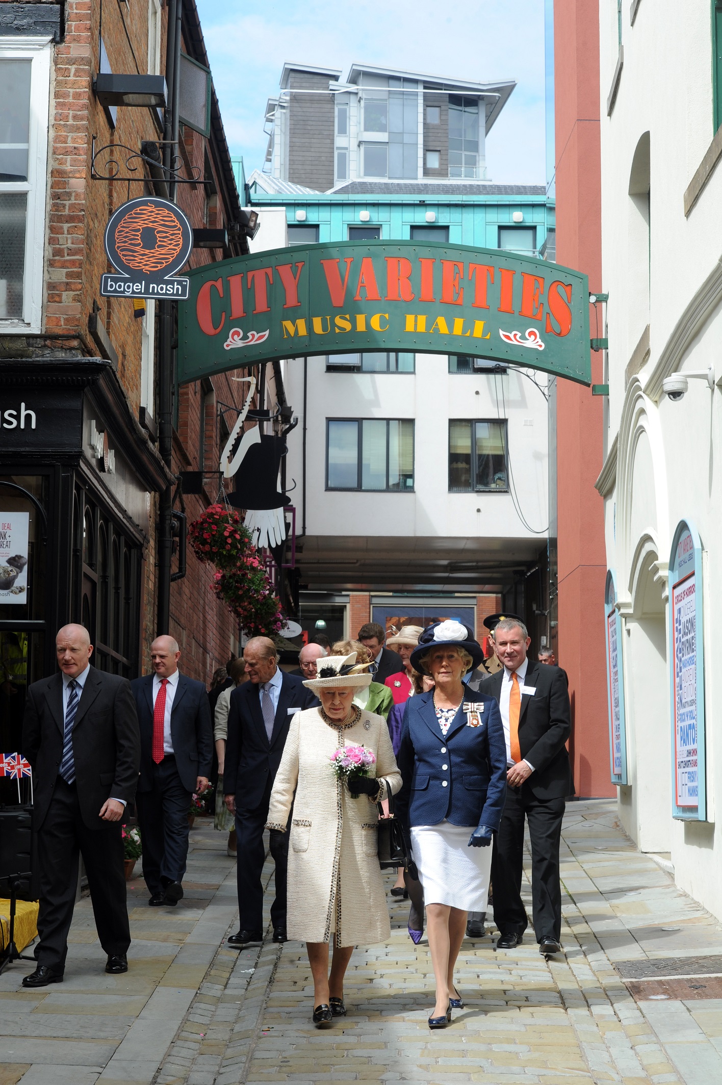 The Queen's Visit The Queen wearing a cream dress and hat followed by a group of people in suits walked down a narrow road. The sign above her reads City Varieties Music Hall.