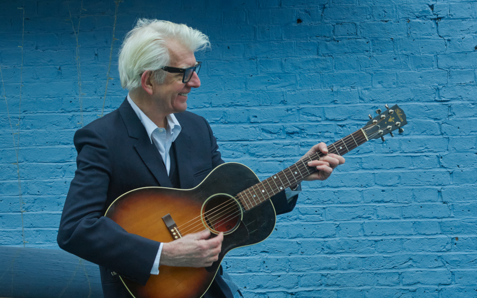 Nick Lowe Nick Lowe playing the acoustic guitar against a blue brick wall
