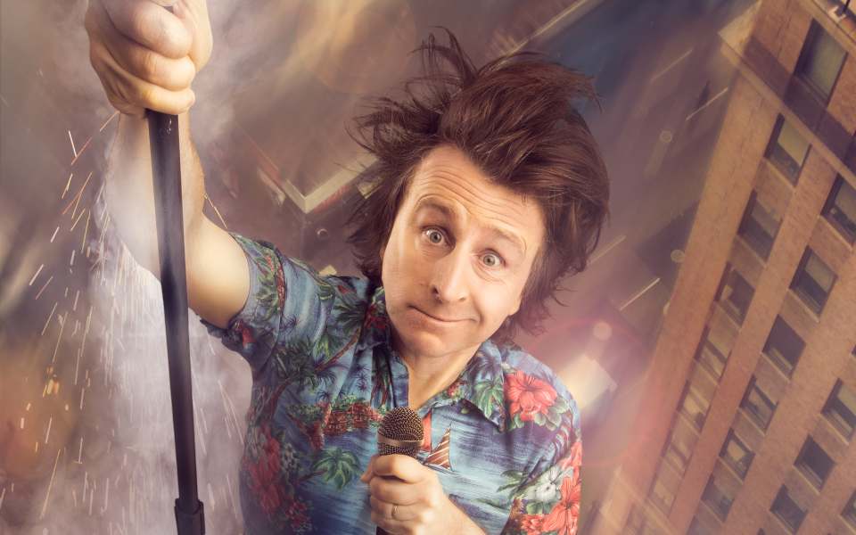 Milton Jones Milton Jones wearing a hawaiian shirt, messy brown hair, holding a microphone