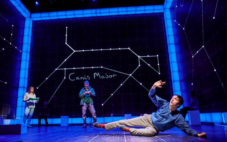 Connor Curren (Christopher) in The Curious Incident of the Dog in the Night-Time A boy in a blue hoodie lays at the front of the stage surrounding by black screens showing images of constellations