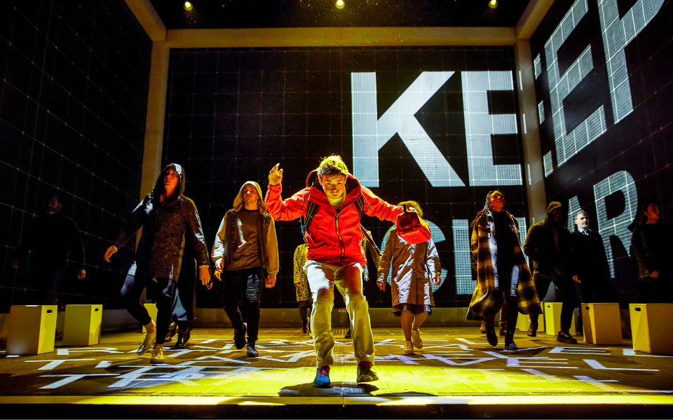 The Curious Incident of the Dog in the Night-Time A boy in a red hoodie stands at the front of the stage surrounded by people
