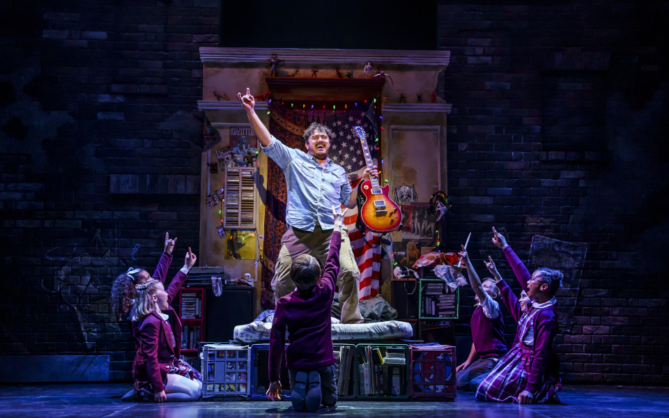 School of Rock Jake Sharp playing Dewey Finn standing on a bed with a guitar surrounded by kids kneeling around him