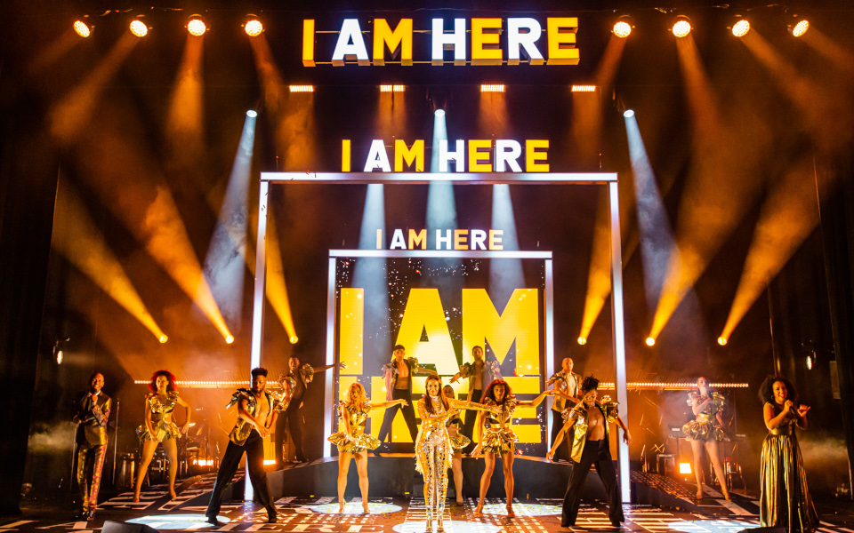 Oti Mabuse I Am Here Oti Mabuse and her dancers on stage in front of big gold and white letters says I Am Here four times