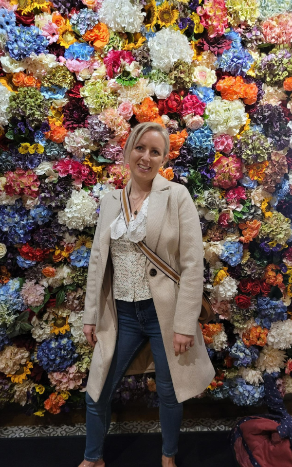 Kirsty Triplett Kirsty Triplett wearing jeans and white shirt smiles to camera against a background of brightly coloured flowers