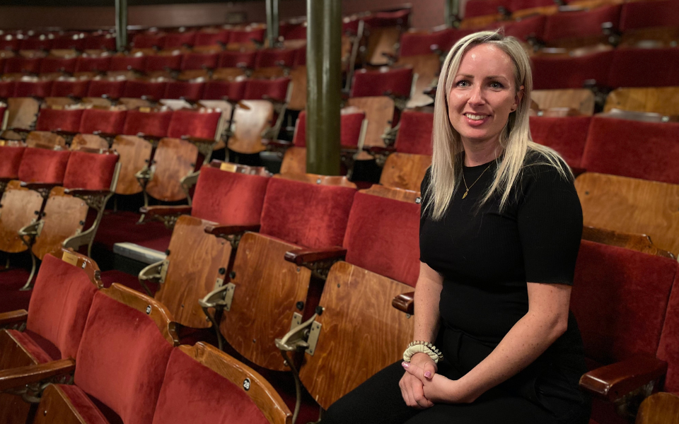 Kirsty Triplett Kirsty Triplett, wearing a black t-shirt and trousers, smiles to camera whilst sat in the auditorium at Leeds Grand Theatre