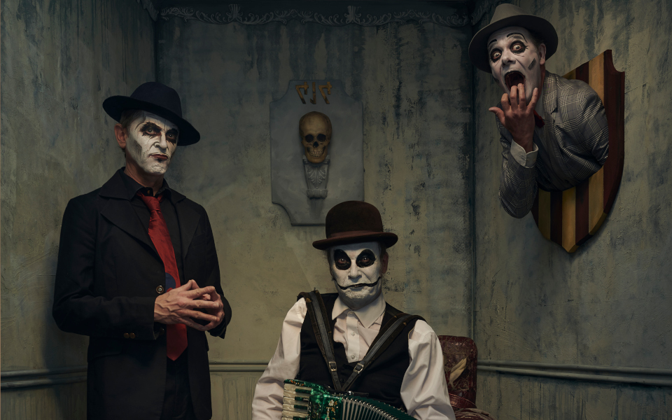 The Tiger Lillies The Tiger Lillies wearing their iconic white make-up and sat in a creepy dark room, one member has their head hanging on the wall like a hunters trophy, another is sat on a chair playing an accordion