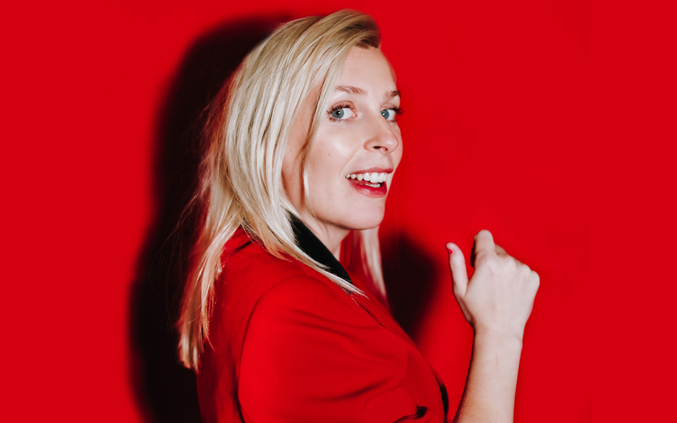 Sara Pascoe Sara Pascoe wearing a red blazer and red lipstick, smiling against a red background